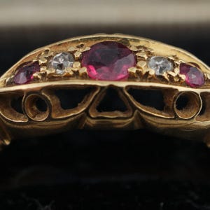 May include: A gold ring with a delicate design featuring five red and white gemstones. The ring has a detailed, intricate design.