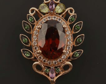 May include: A rose-coloured metal pendant featuring a large oval dark red central gemstone surrounded by a halo of small clear stones. The frame is decorated with marquise-cut green and purple stones, with a dangling purple teardrop gemstone.