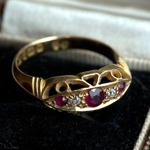 May include: A gold ring featuring a central round-cut ruby flanked by two smaller round-cut diamonds and two additional smaller rubies on the outer edges. The band has an openwork design with decorative metal scrollwork along the top section.