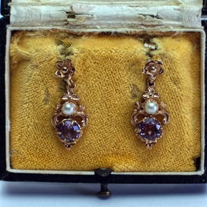 May include: A pair of antique gold-toned earrings displayed in a vintage jewelry box. Each earring features a flower-shaped top, a small pearl, and a large, round, purple gemstone. The earrings are set against a yellow velvet background.