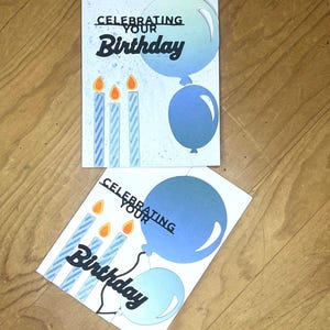 Könnte beinhalten: Zwei Geburtstagskarten mit dem Text "Celebrating Your Birthday". Jede Karte zeigt blaue Luftballons und Kerzen mit orangefarbenen Flammen. Die Karten liegen auf einer Holzoberfläche.