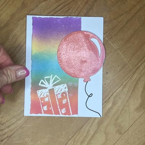Könnte beinhalten: Eine handgefertigte Grußkarte mit einem farbenfrohen Hintergrundverlauf aus Lila, Blau und Orange. Auf der Karte sind ein rosa Ballon und eine Geschenkbox abgebildet. Der Ballon hat ein strukturiertes Aussehen und ist an einer schwarzen Schnur befestigt.