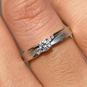 Puede incluir: Un anillo de plata con una piedra preciosa central redonda y transparente, flanqueada por piedras transparentes más pequeñas. La banda del anillo es lisa y pulida, con una ligera curva. El anillo se muestra en un dedo.
