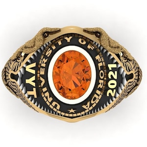 May include: A gold-colored ring with a central orange gemstone. The ring features two alligator heads on either side, with the text "UNIVERSITY OF FLORIDA 2021" around the gemstone. The ring has a black and gold color scheme.