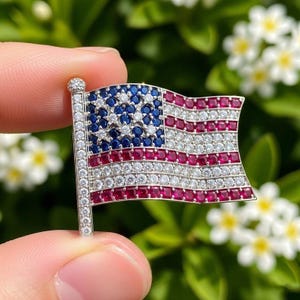 May include: A brooch shaped like the American flag, featuring a silver-tone metal frame. The flag design is embellished with blue, red, and white gemstones, representing the stars and stripes of the US flag.
