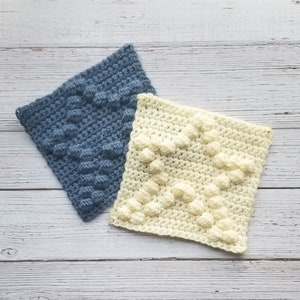 May include: Two crocheted squares, one in a light blue color and one in a cream color, with a textured, raised pattern. The squares are on a white wooden surface.