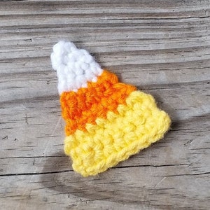 May include: A handmade crochet candy corn in white, orange, and yellow. The candy corn is made with a textured stitch and is sitting on a weathered wooden surface. The colors are bright and the craft is well-executed.