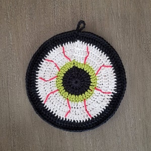 Puede incluir: Un posavasos de ganchillo con forma de ojo en un esquema de color negro, blanco, verde y rojo.