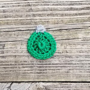May include: A green crocheted Christmas bauble with a white top. The bauble is on a wooden background.