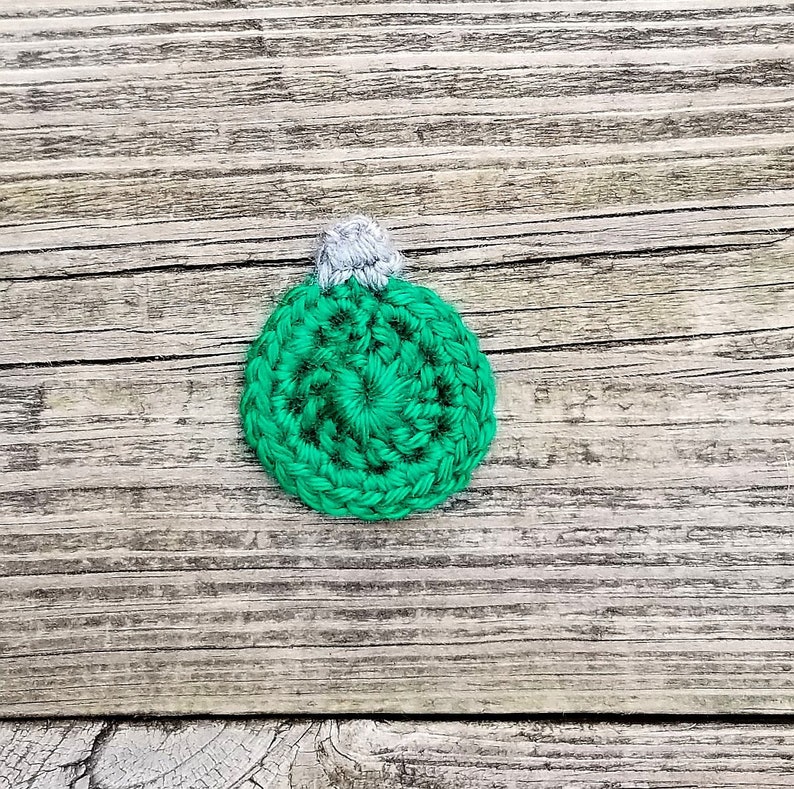 May include: A green crocheted Christmas bauble with a white top. The bauble is on a wooden background.