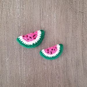 May include: Two crocheted watermelon slices. Each slice features a pink center with black seed details, a white inner rind, and a green outer rind. The slices are arranged on a wooden surface.