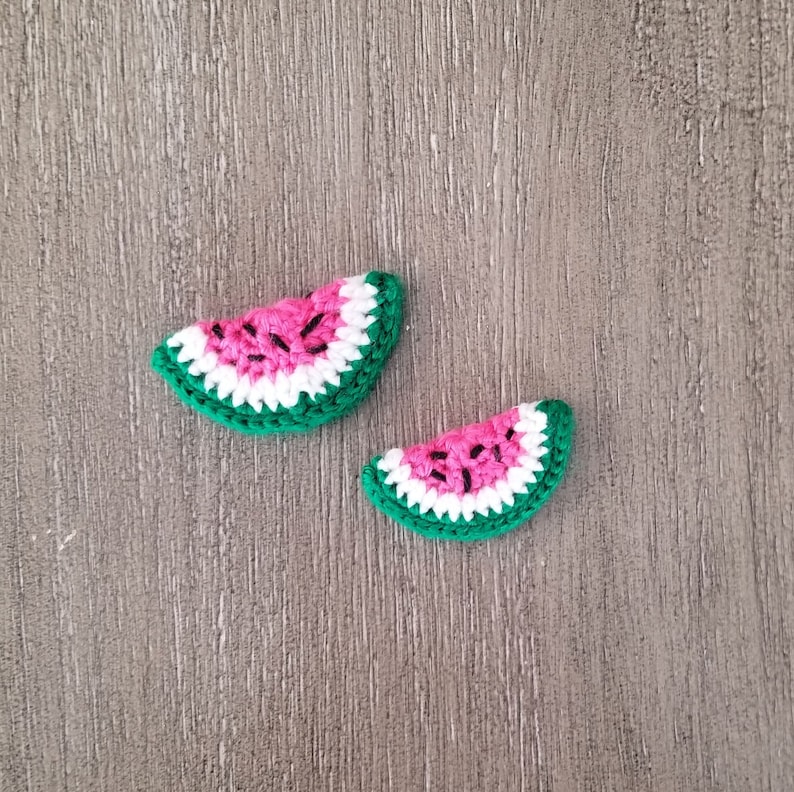 May include: Two crocheted watermelon slices. Each slice features a pink center with black seed details, a white inner rind, and a green outer rind. The slices are arranged on a wooden surface.