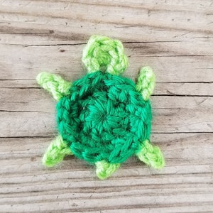 K&ouml;nnte beinhalten: Eine gr&uuml;ne geh&auml;kelte Schildkr&ouml;te mit einem hellgr&uuml;nen Panzer und Kopf. Die Schildkr&ouml;te sitzt auf einer Holzoberfl&auml;che.