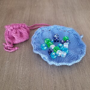 Pode incluir: Uma bolsa de dados de crochê azul com uma bolsa de cordão de crochê rosa no fundo. A bolsa azul está cheia de dados azuis, verdes e brancos.