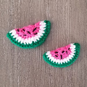May include: Two handmade crochet watermelon slices. Each slice features a vibrant pink center with black seed details, bordered by a white rind and a green outer edge. The craftwork is displayed on a wooden surface, showcasing the texture and detail of the yarn.