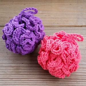 Può includere: Due pouf da bagno all'uncinetto, uno viola e uno rosa, su una superficie di legno.