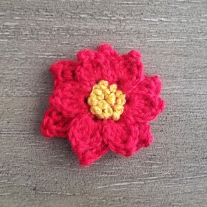 May include: A crocheted red flower with a yellow centre. The flower is made with yarn and has a textured appearance.