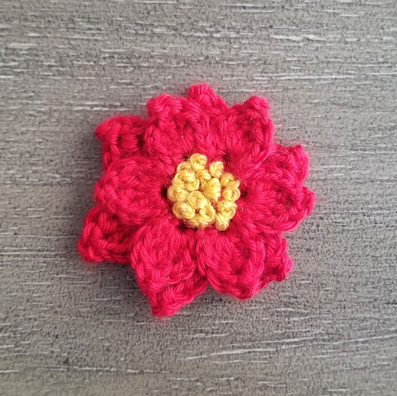 May include: A crocheted red flower with a yellow centre. The flower is made with yarn and has a textured appearance.