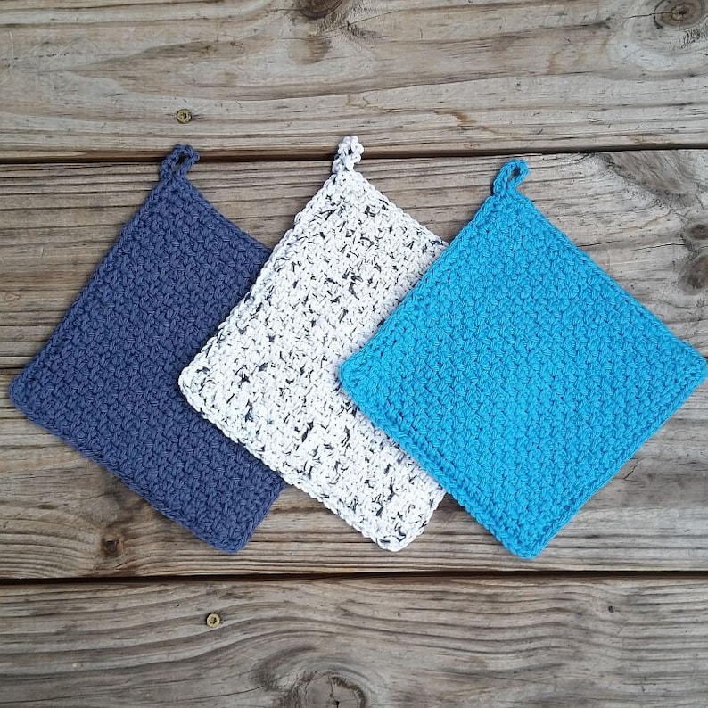 May include: Three crocheted pot holders in different colors: blue, white with black flecks, and gray. The pot holders are square shaped and have a loop at the top for hanging.