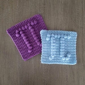 Könnte beinhalten: Zwei gehäkelte Quadrate, eines lila und eines hellblau, mit einem erhabenen Texturmuster. Die Quadrate sind mit einer Noppenmasche gearbeitet.