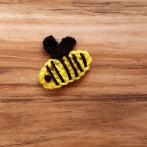 Peut inclure: Une abeille en crochet avec des ailes noires et des rayures jaunes et noires. L'abeille est sur une surface en bois.