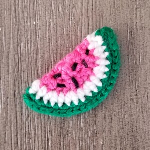 May include: A crocheted watermelon slice featuring a pink center with black seeds, white rind, and a green outer edge. The handmade craft item is likely a decorative piece or an accessory.