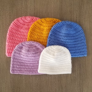 May include: A selection of six crocheted beanies in shades of pink, yellow, blue, purple, and white. The hats are arranged in a semi-circle, highlighting their handmade texture and bright colours. The beanies are made of soft yarn.
