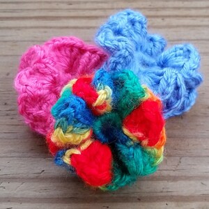 May include: A colorful crocheted flower with red, blue, yellow, and green yarn. The flower is surrounded by two crocheted pieces, one pink and one blue.