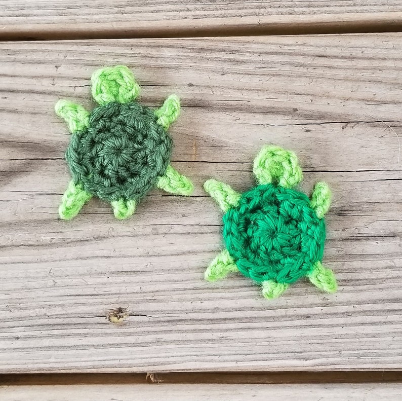 K&ouml;nnte beinhalten: Zwei geh&auml;kelte gr&uuml;ne und hellgr&uuml;ne Schildkr&ouml;ten-Ornamente auf einer Holzoberfl&auml;che.