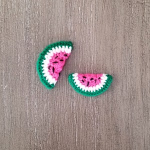 May include: Two crochet watermelon slices. The slices have a bright pink center with black seed details, a white rind, and a green outer edge. The items are handmade and likely used for crafting or embellishment.