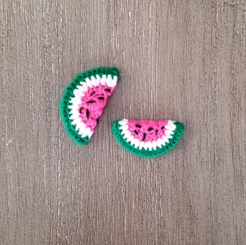 May include: Two crochet watermelon slices. The slices have a bright pink center with black seed details, a white rind, and a green outer edge. The items are handmade and likely used for crafting or embellishment.