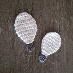 May include: Two crocheted lightbulb ornaments in white with gray bases. The ornaments are on a wood surface.