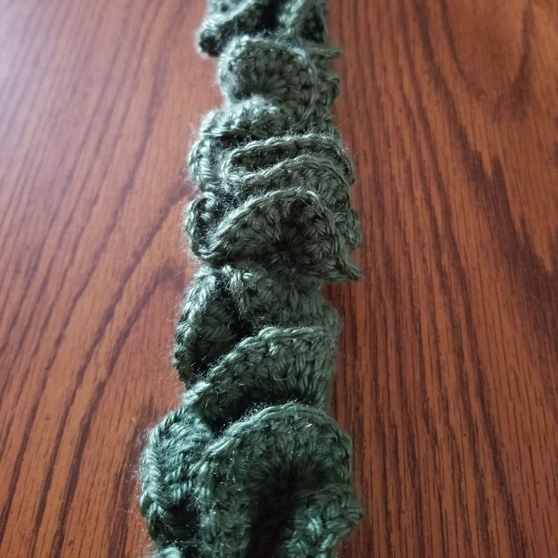 Pu&ograve; includere: Primo piano di una ghirlanda di uncinetto verde con una texture arricciata. La ghirlanda &egrave; fatta di filato e ha un aspetto morbido e peloso.