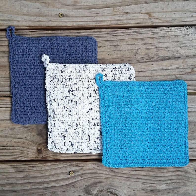 May include: Three crocheted pot holders in different colors: blue, white with black flecks, and light blue. The pot holders are square and have a loop for hanging.