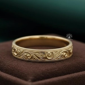 May include: A gold-colored ring with an intricate floral pattern. The ring is set on a dark brown surface, with a green and dark green background. The ring's design features detailed swirls and leaf-like shapes.