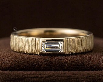 May include: A gold ring featuring a textured, bark-like finish on the band and a single, horizontally set baguette-cut clear gemstone centered on the front. The metal has a warm, metallic yellow tone and a matte, organic surface texture.