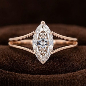 May include: A rose gold ring featuring a large, marquise-cut clear gemstone set in a prong setting. The band is split into two thin, smooth metal arms that meet at the base of the stone, creating a sleek, open design with a polished finish.