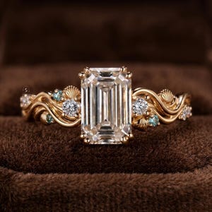 May include: A gold ring featuring a large emerald-cut centre stone, flanked by smaller round clear stones and light blue accent stones. The band has a wavy, organic design with small shell-like details integrated into the metalwork.