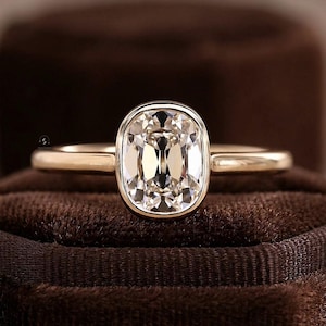 May include: A gold ring featuring a central cushion-cut gemstone set in a smooth bezel setting. The band is polished gold with a rounded profile, showcasing a clear, faceted stone that reflects light with multiple internal facets and sharp edges.