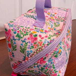 Könnte beinhalten: Rechteckige Tasche mit floralem Muster, mit lavendelfarbenem Reißverschluss und Griff. Die Tasche zeigt ein sich wiederholendes Muster aus Blumen und Schmetterlingen in Rosa, Lila, Grün und Gelb. Reißverschluss und Griff sind aus passendem Lavendelstoff.