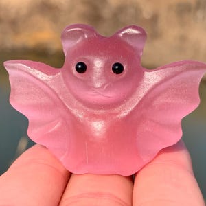 Può includere: Una statuetta a forma di pipistrello rosa con ali spiegate e piccoli occhi neri. Il materiale traslucido lascia passare la luce, creando una morbida lucentezza. Il pipistrello è tenuto in mano, con uno sfondo sfocato di acqua e fogliame.