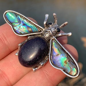 Puede incluir: Un broche de abeja de plata con alas de concha de abulón iridiscente y un cuerpo de piedra azul oscuro.