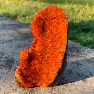 Op de afbeelding: Een grote oranje kristalkluster op een donkerbruine houten basis. De kristalkluster is gemaakt van veel kleine, gefacetteerde kristallen.