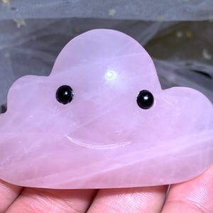 May include: A pink cloud-shaped crystal with black eyes and a carved smile. The crystal is smooth and polished.