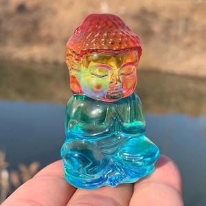 Puede incluir: Una pequeña figurita de vidrio de colores arcoíris de un Buda meditando. La figurita está sentada en posición de loto con las manos juntas frente al pecho.