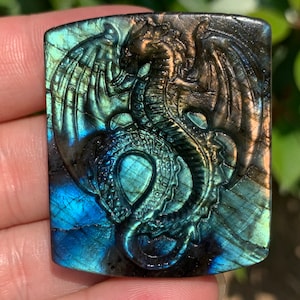 以下が含まれることがあります： 詳細なドラゴンデザインが施された、彫刻されたラブラドライトの宝石ペンダント。ドラゴンは翼を広げ、蛇のような体で描かれています。宝石は青、緑、金色の虹色の輝きを放っています。