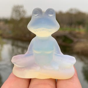 Puede incluir: Una figura de rana translúcida y opalescente en posición sentada. La rana es de color azul claro y amarillo, con una superficie lisa y pulida. La figura mide aproximadamente 5 cm de altura.