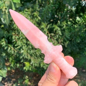 Puede incluir: Una daga de cristal rosa con una hoja puntiaguda y un mango detallado. La daga se sostiene sobre un fondo de follaje verde, mostrando su color y forma. El diseño de la daga sugiere que podría ser un artículo decorativo o de colección.