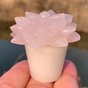 Op de afbeelding: Een roze roos gesneden uit rozenkwarts, in een witte pot. De rozenkwartsbloem is een populaire kristal voor liefde en genezing.