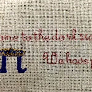 May include: Cross-stitch embroidery on a beige fabric with red thread. The text reads "Come to the dark side. We have pie." A blue and yellow pie with steam coming off of it is stitched below the text.